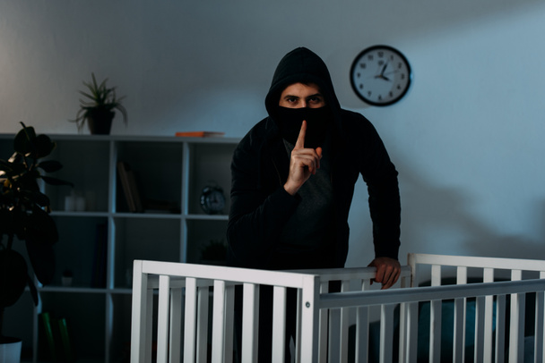 Kidnapper in Maske und schwarzem Kapuzenpullover zeigt Schweigezeichen neben Krippe - Foto, Bild