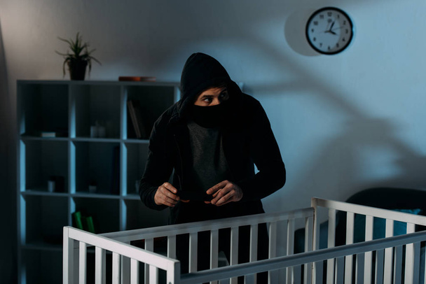 Worried kidnapper in black mask standing near crib and taking picture - Photo, Image