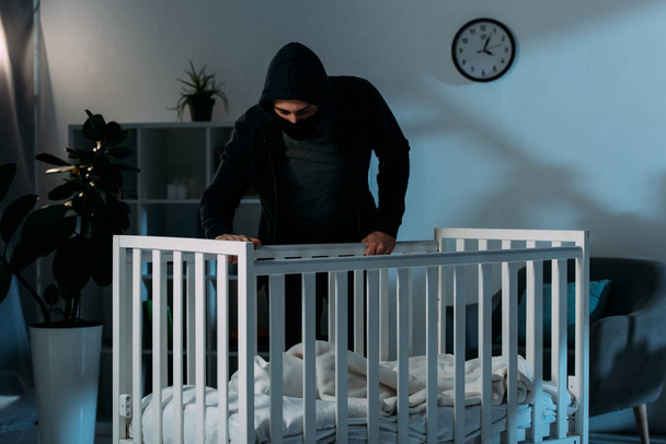 Похититель в черной одежде стоит в темной комнате и смотрит в кроватку
 - Фото, изображение
