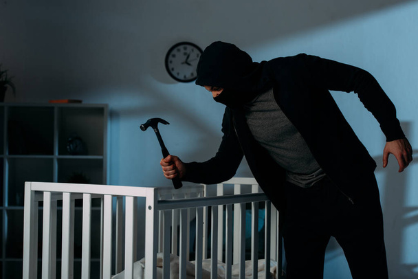 Злочинець у масці тримає молоток, стоячи біля ліжечка в темній кімнаті
 - Фото, зображення