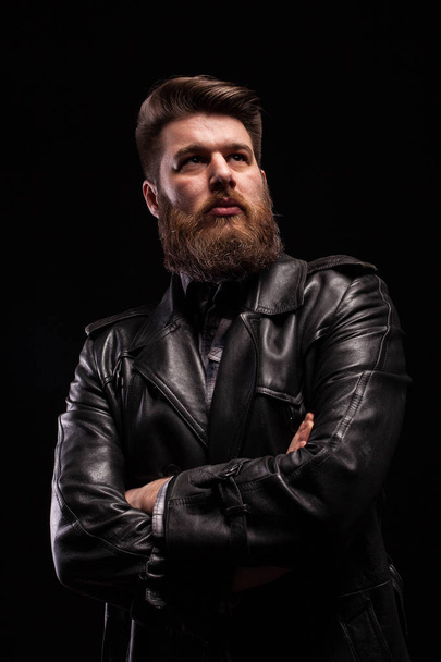 Porträt eines gutaussehenden bärtigen Mannes in einer Lederjacke mit ernstem Ausdruck vor schwarzem Hintergrund - Foto, Bild