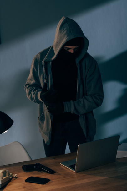 マスクやパーカーの暗い部屋でノート パソコンを使用しての犯罪者 ロイヤリティフリー写真 画像素材