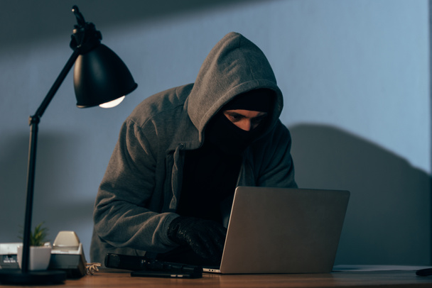 Criminal en máscara y sudadera con capucha usando portátil en habitación oscura
 - Foto, imagen