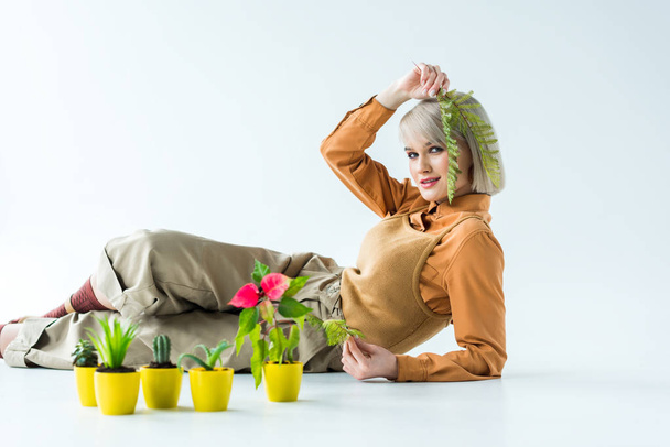 hermosa chica con estilo mirando a la cámara y sosteniendo hojas de helecho mientras posa cerca de macetas aisladas en blanco
 - Foto, Imagen