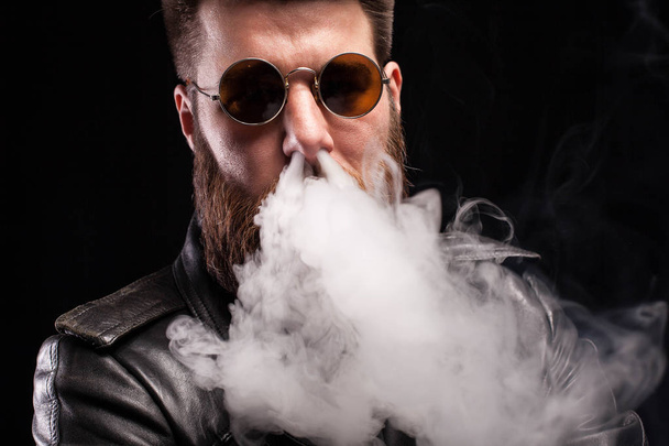 Homem barbudo atraente exalando fumaça de cigarro em seu nariz sobre fundo preto
 - Foto, Imagem