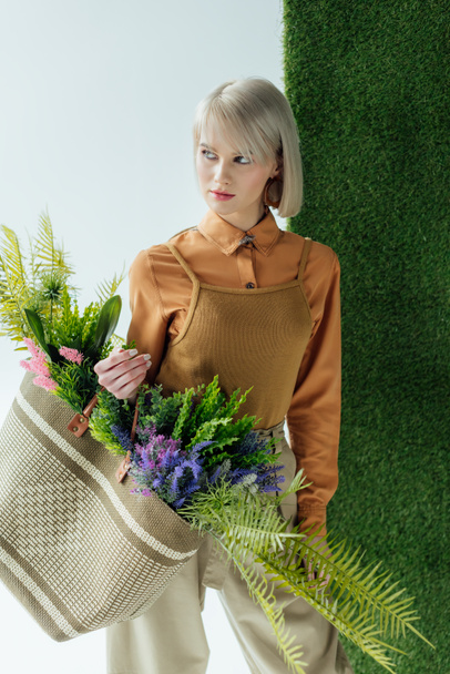 beautiful stylish girl holding bag with fern and flowers on white with green grass  - Фото, изображение