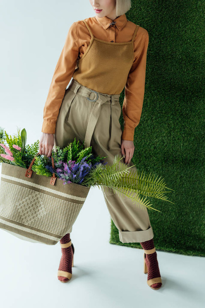 cropped view of stylish young woman holding bag with fern and flowers on white with green grass  - Foto, Imagen