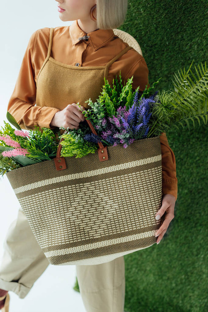 bijgesneden weergave van stijlvolle jongedame bedrijf tas met varens en bloemen in wit met groen gras  - Foto, afbeelding