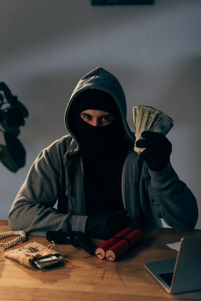 Terrorist in masker en handschoenen aan tafel zit met wapen en bezit dollar biljetten - Foto, afbeelding