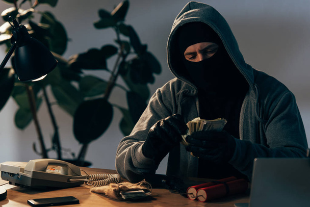 Terrorist in mask and gloves counting dollar banknotes in room - Fotografie, Obrázek
