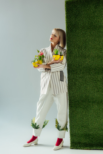 beautiful stylish girl posing while holding flower pots on grey with green grass  - Zdjęcie, obraz