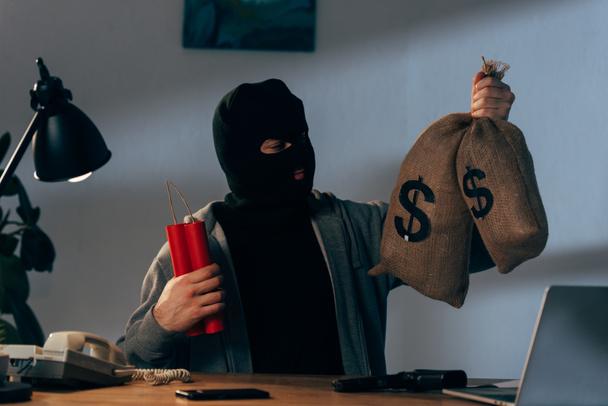 Terrorista en máscara sosteniendo dinamita y mirando bolsas de dinero en la habitación
 - Foto, imagen