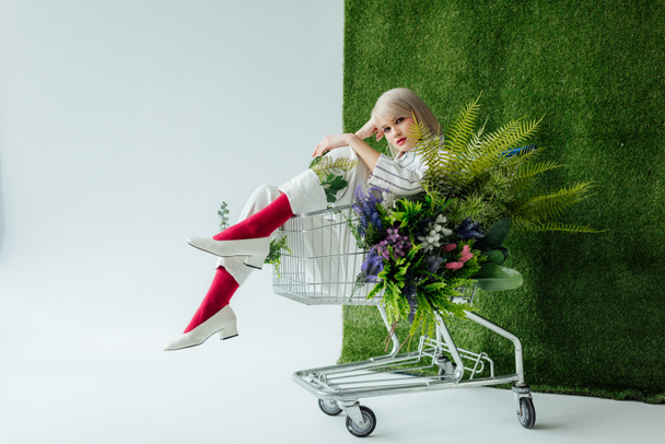 beautiful fashionable girl sitting in shopping cart with fern and flowers on white with green grass - Photo, Image