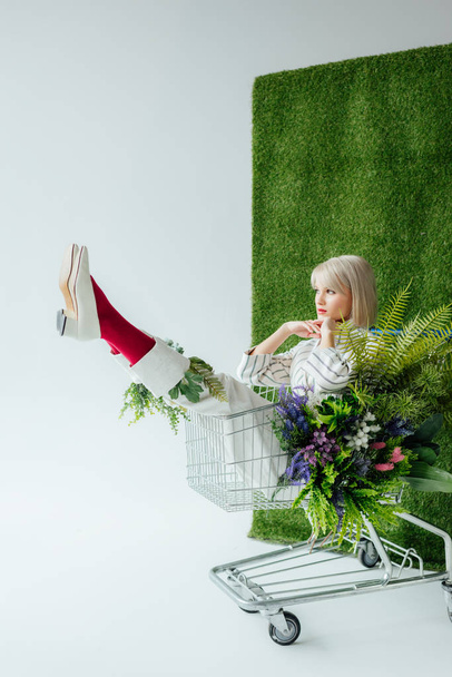 beautiful stylish girl sitting in shopping cart with flowers on white with green grass - Foto, afbeelding