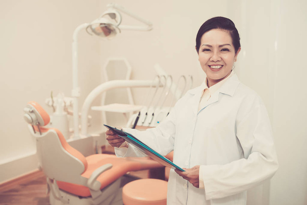 Joyeux asiatique adulte médecin dans le cabinet dentaire tenant presse-papiers et souriant à la caméra
 - Photo, image