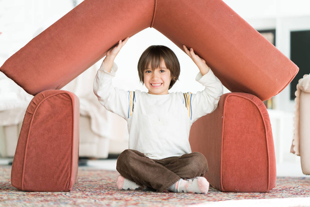 Kleiner Junge mit langen Haaren spielt zu Hause und baut Haus mit Dach - Foto, Bild