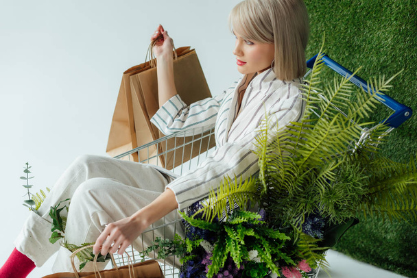 beautiful stylish girl sitting in cart with fern, flowers and shopping bags on white with green grass - Foto, imagen