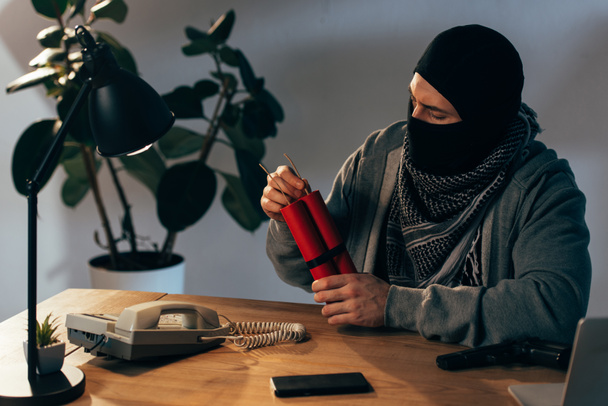 Terrorist in mask sitting at table with dynamite in room - Photo, Image