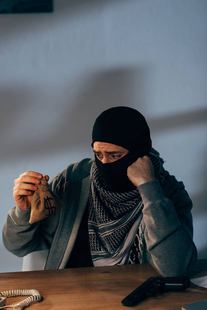 Грустный террорист в маске смотрит на маленькую сумку с деньгами в комнате
 - Фото, изображение