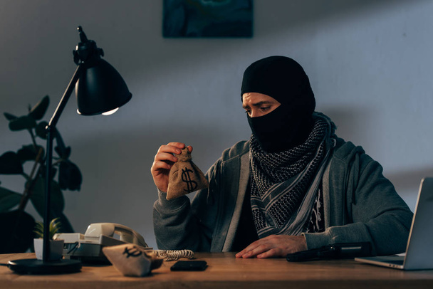 Upset terrorist in mask sitting in room and holding small money bag - Photo, Image