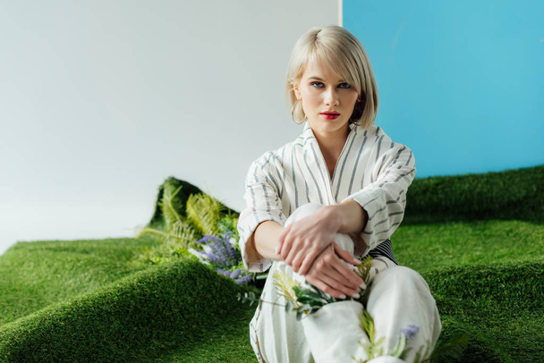 красивая стильная блондинка смотрит в камеру и позирует, сидя на искусственной траве
  - Фото, изображение
