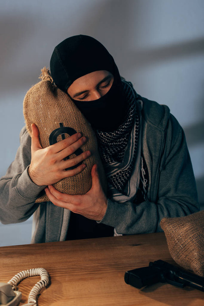 Terrorista satisfeito em máscara abraçando saco de dinheiro com olhos fechados
 - Foto, Imagem