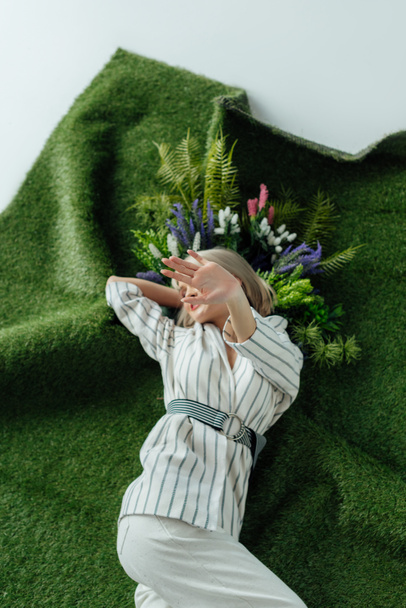 stilvolles Mädchen, das Gesicht mit der Hand bedeckt, während es auf Kunstrasen mit Farn und Blumen liegt - Foto, Bild