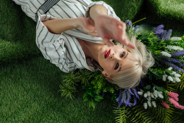 selective focus of beautiful stylish girl with outstretched hands lying on artificial grass with fern and flowers - Foto, immagini