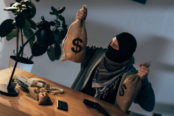 Terrorist in mask sitting at table and holding bags with dollars - Photo, Image