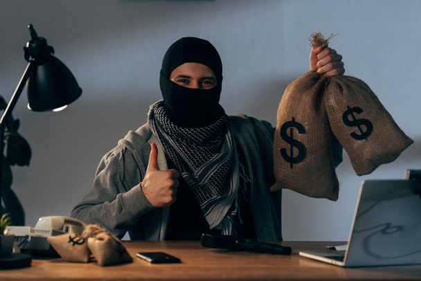 Terrorist in mask holding bags with dollars and showing thumb up - Foto, imagen