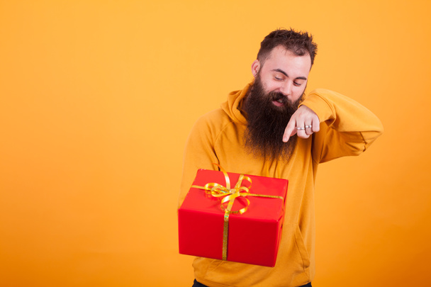 Yakışıklı adam sarı hoodie sarı arka plan üzerinde kırmızı hediye kutusu üzerinde işaret içinde sakallı. - Fotoğraf, Görsel