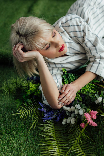 beautiful stylish blonde girl with eyes closed lying on artificial grass with fern and flowers - Photo, Image