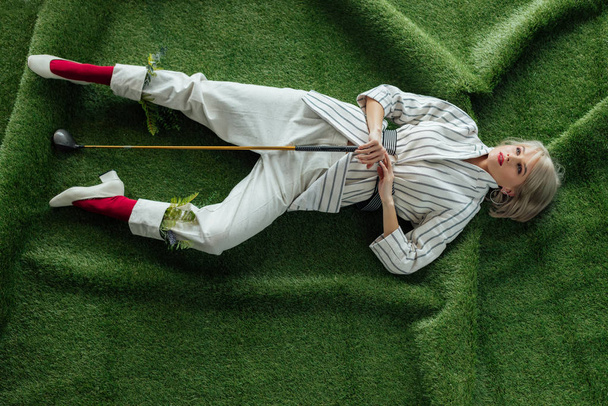 high angle view of beautiful stylish girl lying with golf club on artificial grass - Zdjęcie, obraz
