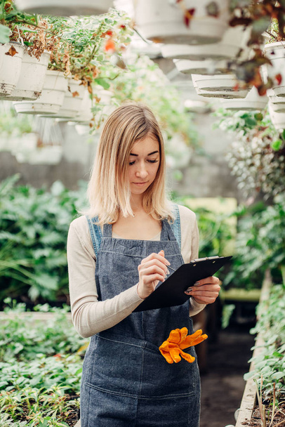 Egy fiatal női kertész feljegyzést vezet a virágokról egy üvegházban.. - Fotó, kép