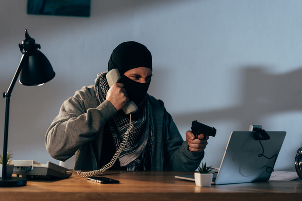 Angry terrorist in mask talking on telephone and showing gun at webcam - Fotografie, Obrázek