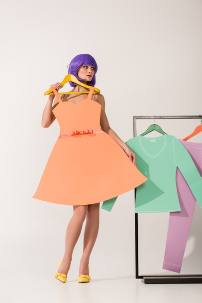 beautiful girl with purple hair holding paper dress and posing near rack with colorful clothes on white  - Photo, Image