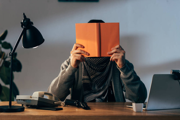 Террорист в шарфе Кеффие с книгой для чтения оружия в комнате
 - Фото, изображение