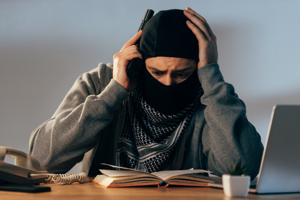 Besorgter Terrorist mit Maske berührt Kopf beim Lesen von Buch - Foto, Bild