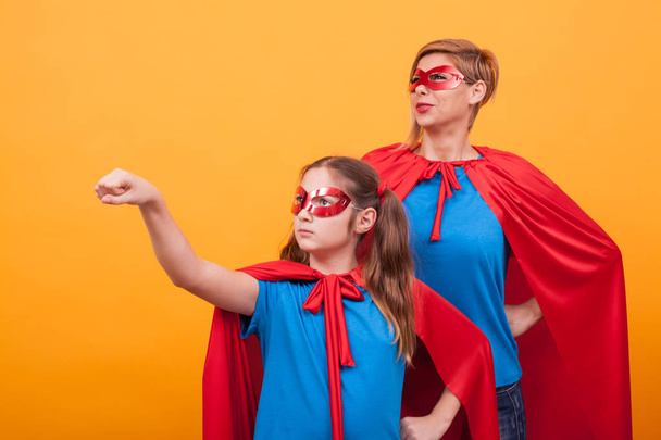 Meisje gekleed als superheros bedrijf in haar vuist in de lucht met haar moeder over eyellow achtergrond. - Foto, afbeelding