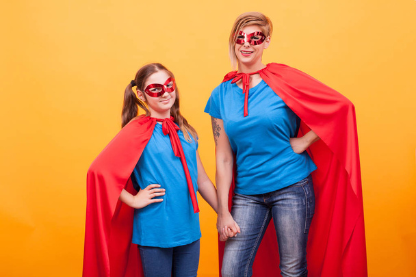 Little girl and mother dressed like superheros holding hands and smiling to the camera over yellow background - Photo, Image