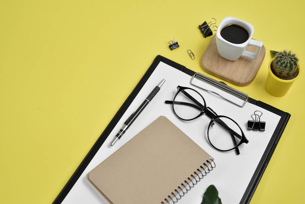 Office desk workspace with blank clip board, office supplies, pen, cactus, green leaf, coffee cup on a wooden stand and eye glasses on yellow background. Flat lay, top view blog concept - 写真・画像