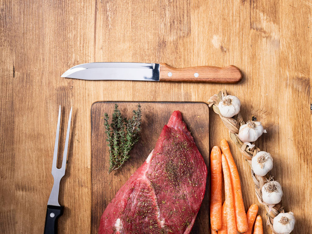 Vue de dessus d'un gros morceau de viande rouge à côté de l'ail, des carottes et du couteau de chef au centre du cadre
 - Photo, image