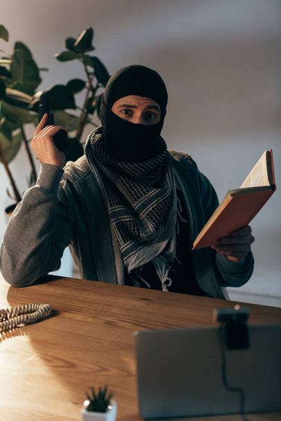 Terrorista con máscara negra sosteniendo pistola y leyendo libro
 - Foto, imagen