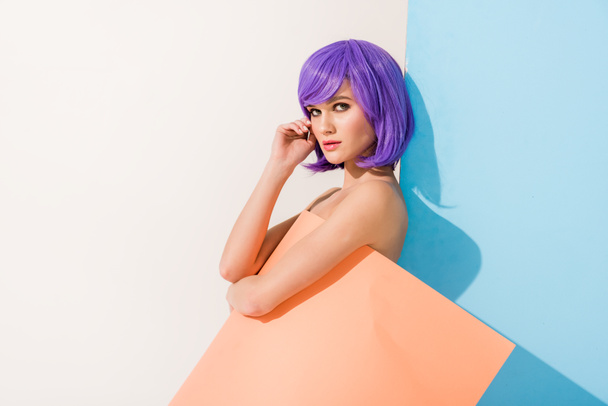 beautiful girl with purple hair covered in coral paper sheet looking at camera while posing on blue and white - Foto, afbeelding