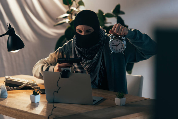 Aggressive terrorist in mask holding gun and showing alarm clock at webcam - Photo, Image