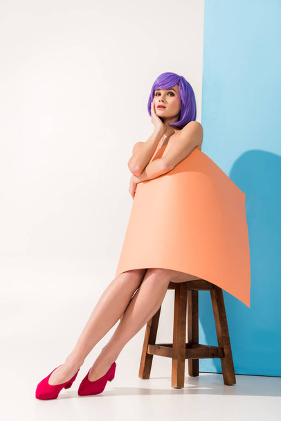 beautiful girl with purple hair covered in coral paper sheet sitting on chair while posing on blue and white - Foto, imagen