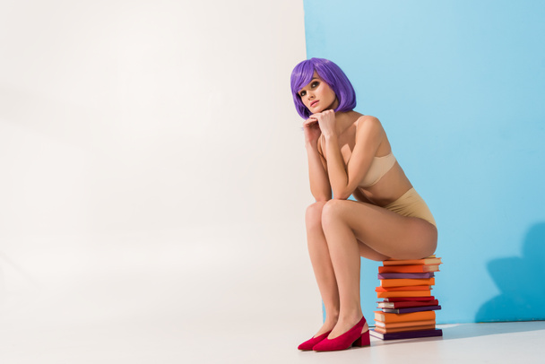 beautiful girl with purple hair looking at camera and sitting on pile of colorful books on blue and white with copy space - Photo, Image