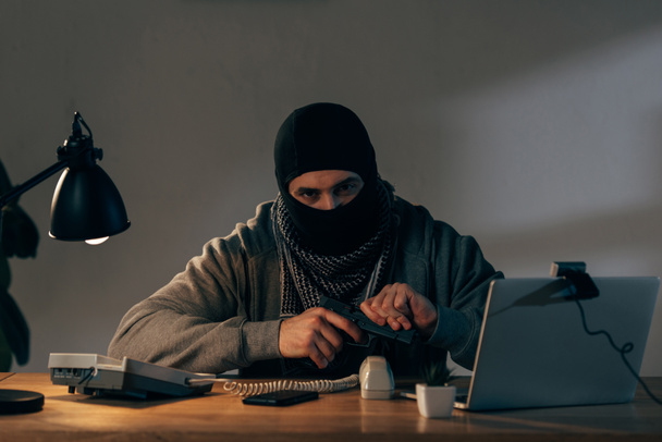 Terrorista con máscara negra sentado a la mesa y cargando pistola
 - Foto, imagen