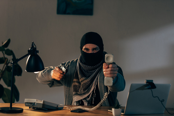 Terrorista aggressivo in maschera che tiene il portatile e mira pistola
 - Foto, immagini