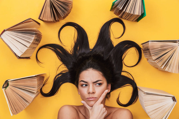Serious young woman lies isolated on yellow background over books. - Φωτογραφία, εικόνα